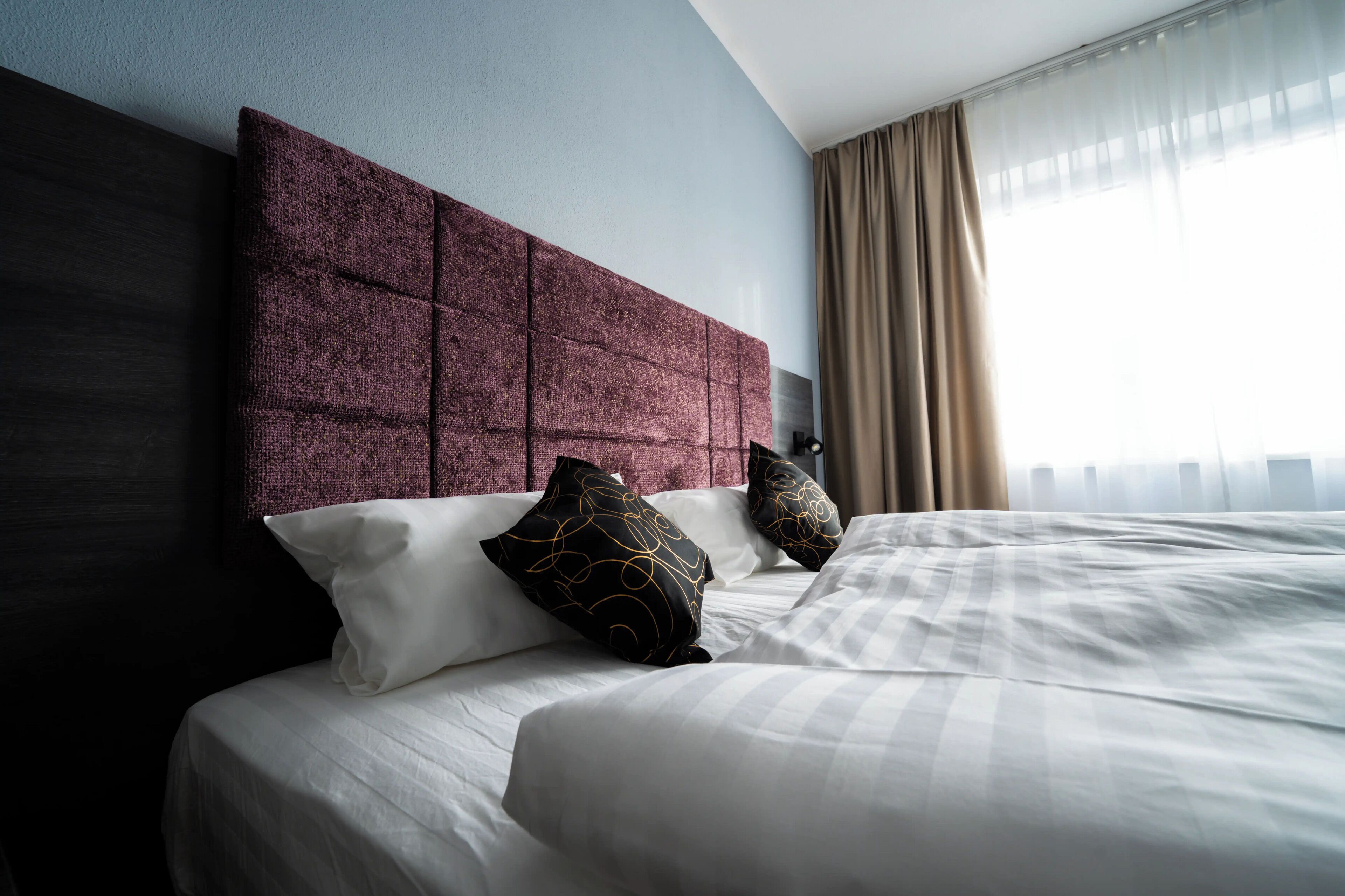 A neatly made bed with white linens and decorative pillows, a dark headboard, and a large window with sheer curtains.
