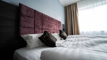Een net opgemaakt bed met witte lakens en decoratieve kussens, een donker hoofdbord en een groot raam met dunne gordijnen.