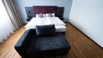 Ein modernes Schlafzimmer mit Bett, einer dunkelgrauen Couch, Holzboden und einem Kopfteil mit lilafarbenen Akzenten.