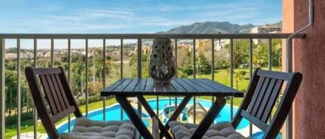 A balcony with two wooden chairs and a table, overlooking a swimming pool and a landscaped area with mountains in the background.