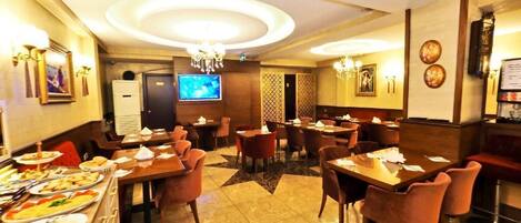 A restaurant with multiple dining tables and chairs, a buffet area with various dishes, and a television screen on the wall.