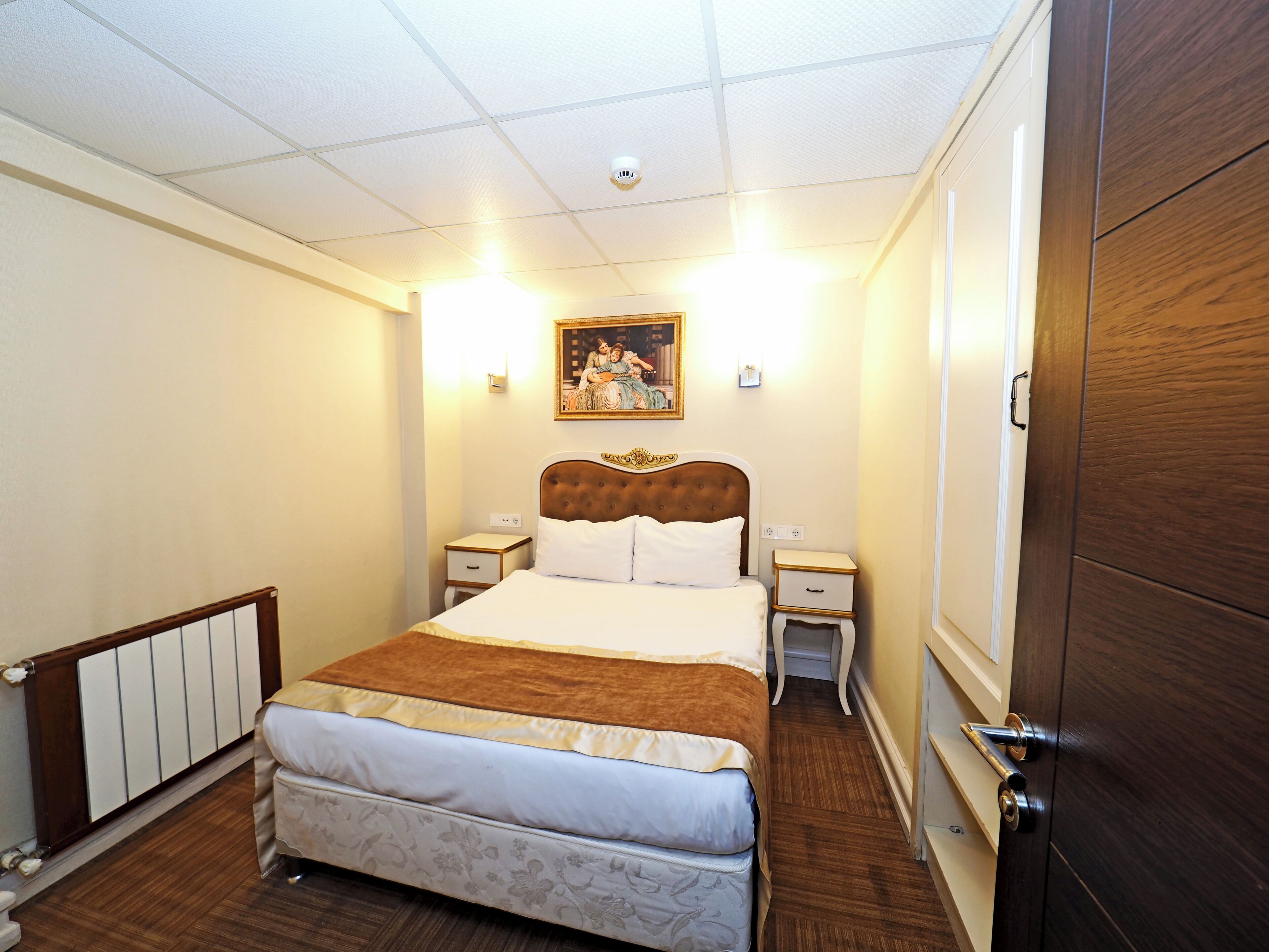 A hotel room with a large bed, a wooden wardrobe, a nightstand, a bedside table, and a painting on the wall.