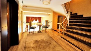 A grand staircase leading to an upper floor, a seating area with a table and chairs, and a view of a room with a large window and curtains. - ELYSİON HOTEL (İstanbul)