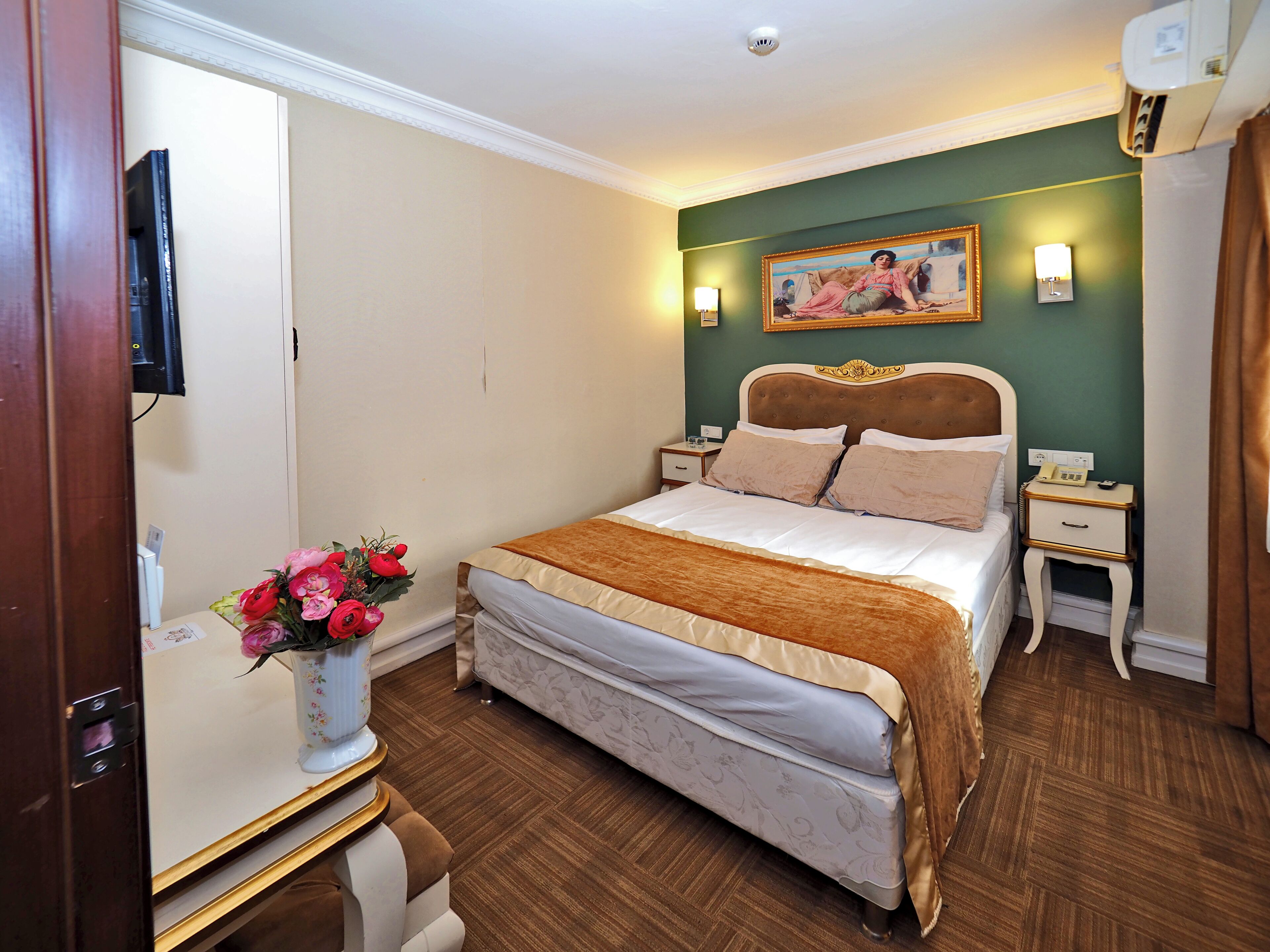 A hotel room with a bed, bedside tables, a nightstand, a wall-mounted TV, a painting, and a vase with flowers.