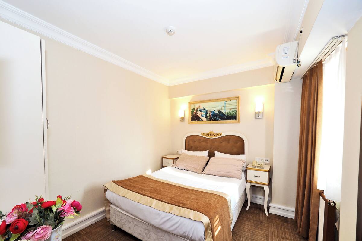 A hotel room with a bed, bedside tables, a nightstand, a vase of flowers, and an air conditioning unit.