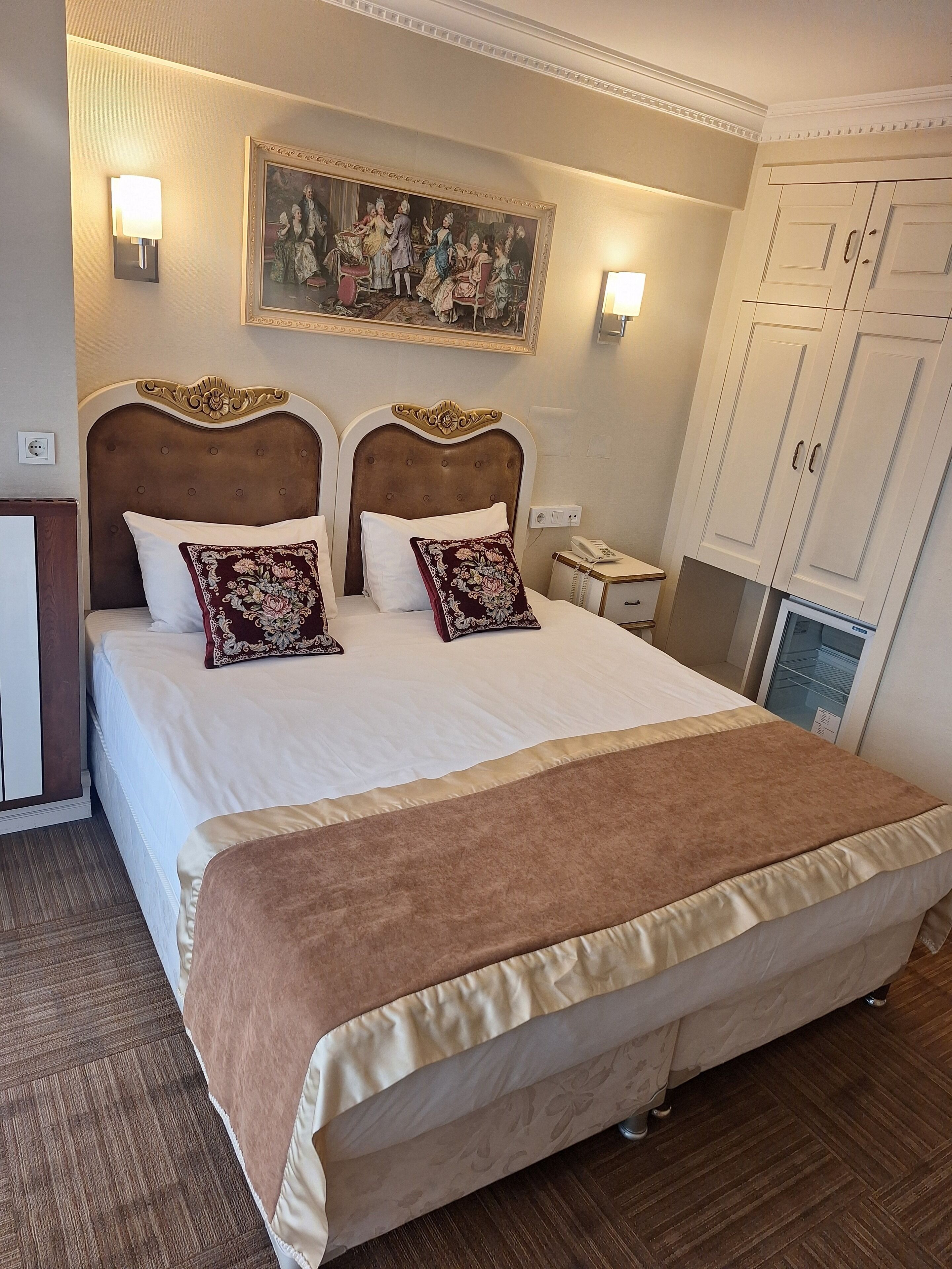 A hotel room with a large bed, two wall-mounted lamps, a painting above the bed, and a wardrobe with double doors.