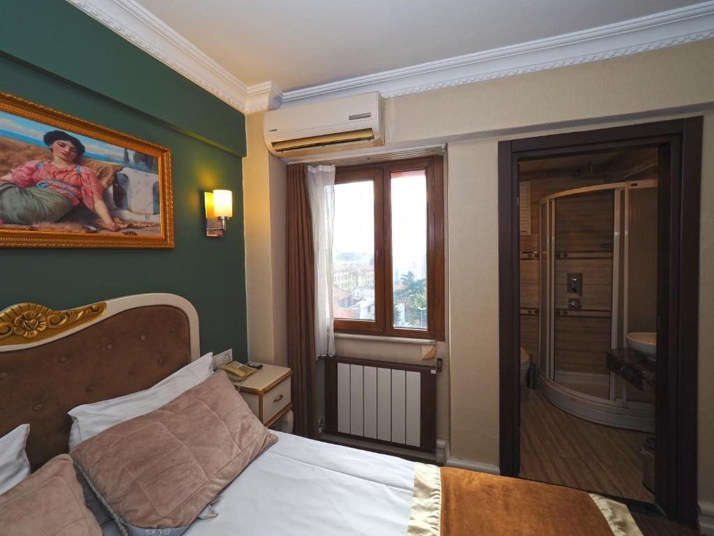 Habitación de hotel con cama, un cuadro en la pared, unidad de aire acondicionado, ventana con cortinas y baño visible a través de una puerta abierta.