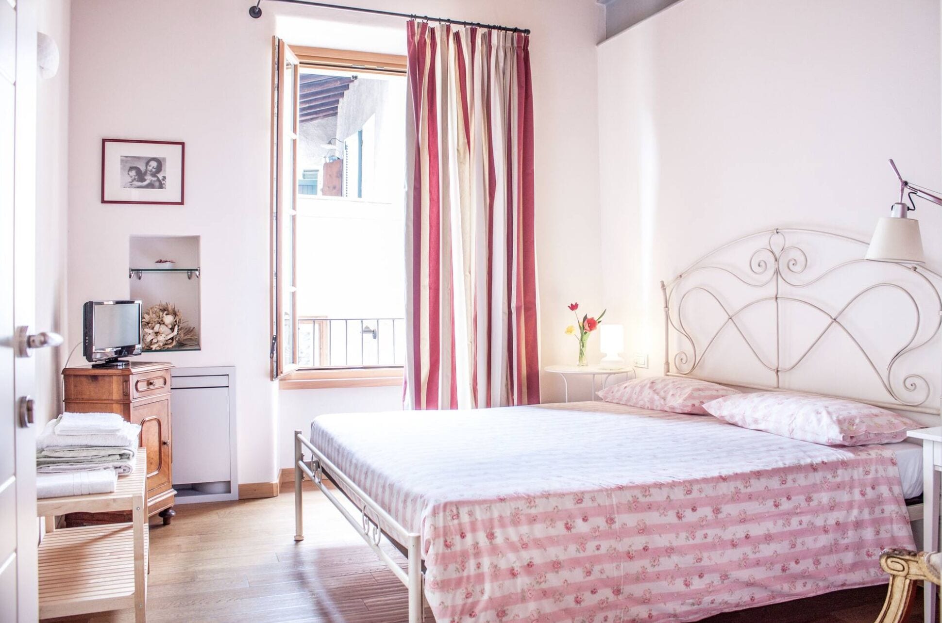 Une chambre à coucher avec un lit, une table de chevet avec une lampe, une commode, un téléviseur posé sur une étagère et une fenêtre avec des rideaux.