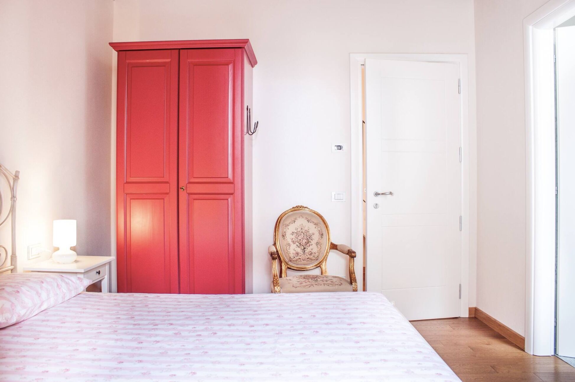 Quarto com cama, guarda-roupa vermelho, porta branca e uma cadeira.