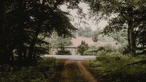 Maalaismaisessa ympäristössä sijaitseva talo, jonka punainen katto erottuu vehreän kasvillisuuden ja puiden keskeltä.
