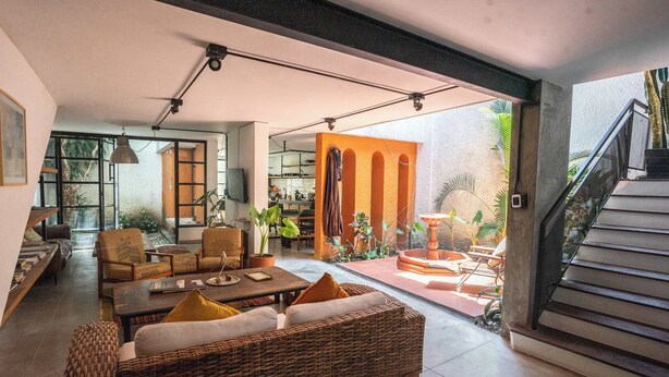 A spacious living area with a large wicker sofa, a coffee table, and a dining table. There is a staircase leading to an outdoor area with plants and a dining set. - w Bright Colonial 5BR House With Balcony (Medellín)