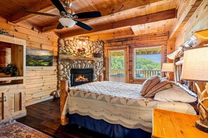 A log cabin bedroom with a stone fireplace, a bed with a patterned bedspread, and a ceiling fan. - 3BR Resort Cabin w/Views! Hot Tub*Games*Comm Pool (Sevierville)