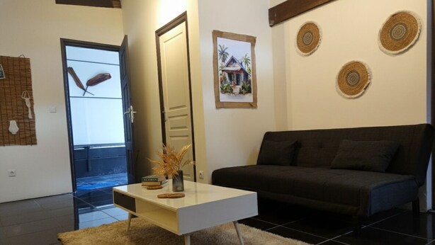 Una sala moderna con una mesa de centro blanca, un sofá negro y un tapete beige. Se observan tres adornos de pared tejidos y un cuadro enmarcado de una casa.