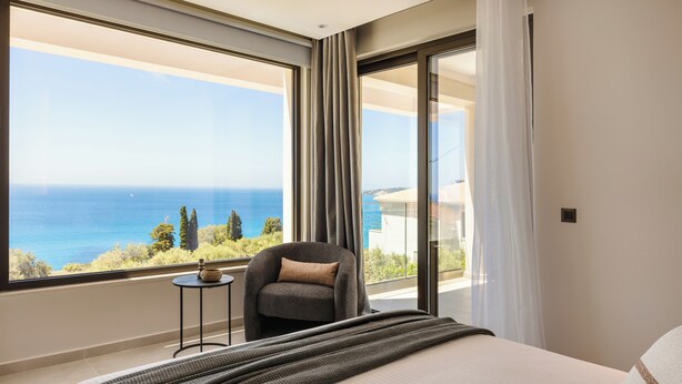 Une chambre dotée d’une grande fenêtre donnant sur la mer, avec un lit, une chaise et une petite table.
