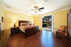 A bedroom with a large bed, a ceiling fan, a nightstand, a chair, and a sliding glass door leading to a patio. - Experience the Florida dream at Villa Barbara in Cape Coral! (Cape Coral)