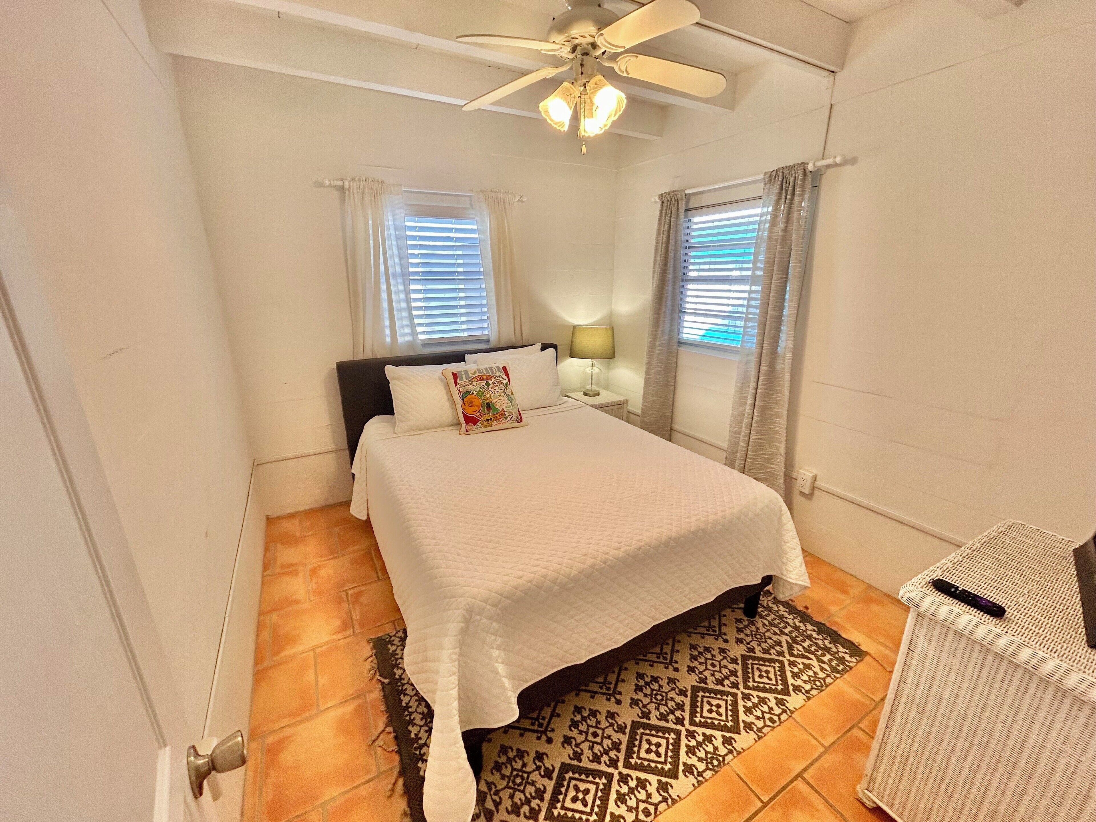 Une chambre à coucher avec un lit, un ventilateur de plafond, une table de chevet avec une lampe et une fenêtre avec des rideaux.