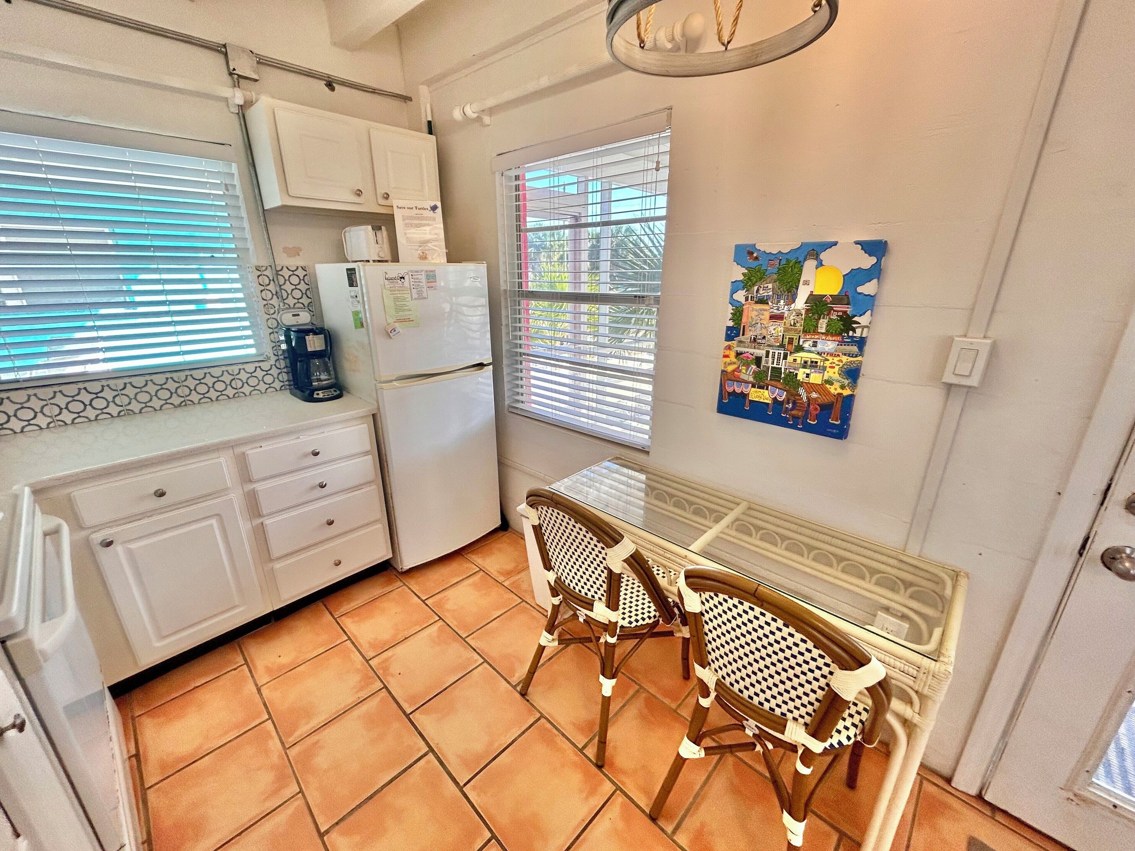 A compact kitchen with white cabinets, a refrigerator, a coffee maker, and a dining area with two wicker chairs.