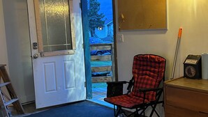 Una habitación con piso de madera, un sillón rojo a cuadros, un aparador de madera, una puerta con panel de vidrio esmerilado, una lámpara de pared y vista a un barrio residencial.