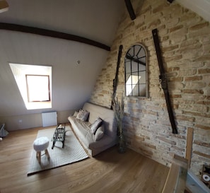 A loft-style room with a sloped ceiling, exposed brick walls, a wooden floor, a sofa with cushions, a small table, and a large window.