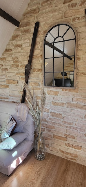 A room with a stone wall, a window with a metal frame, a wooden floor, a sofa with cushions, and a vase with dried plants.