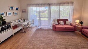 Un espacio habitable pequeño con kitchenette, zona de comedor, sofá y un gran ventanal con cortinas translúcidas.