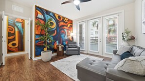 Ein modernes Wohnzimmer mit Holzböden, einem grauen Sofa, einem grauen Sessel und einem farbenfrohen, abstrakten Gemälde an der Wand.