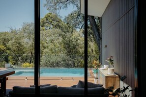 Un área de piscina moderna al aire libre con vista a árboles densos, una terraza de madera y un espacio de descanso con cojines.