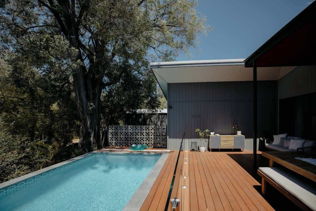 A modern outdoor pool area with a wooden deck, a built-in seating area, and a large tree in the background.