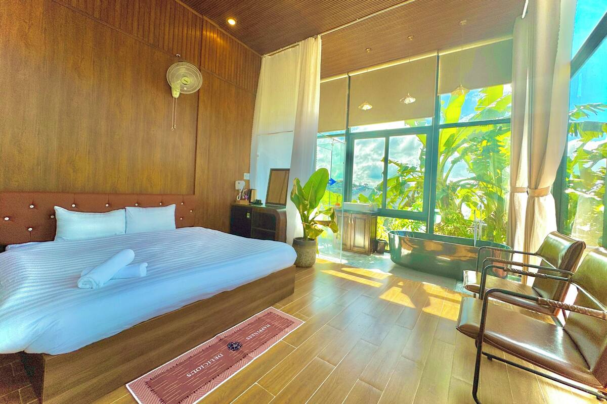 Habitación de hotel con una cama grande, paredes y piso de madera, un escritorio con silla, una planta en maceta y vistas a zonas verdes a través de amplios ventanales.