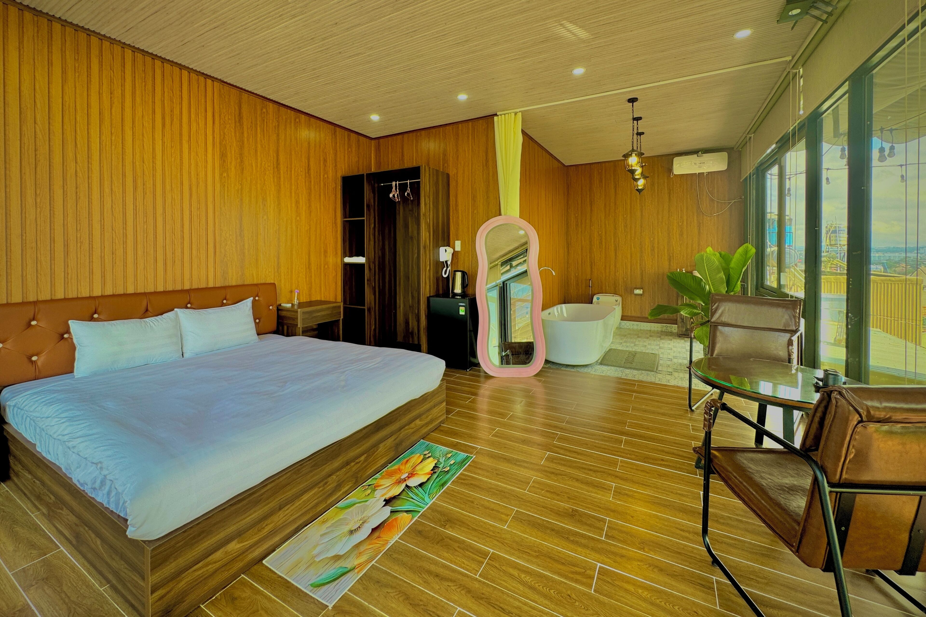A modern hotel room with a large bed, a wooden floor, a glass table with chairs, and a view of the city through large windows.