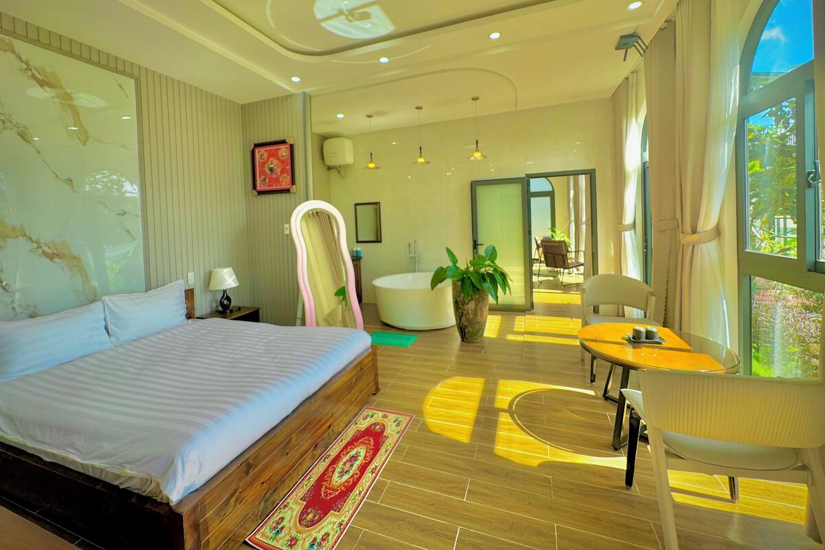 Un dormitorio moderno con una cama grande, una zona de estar con una mesa y sillas, una bañera y un amplio ventanal con cortinas.