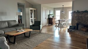Un salón amplio con suelo de madera, un sofá gris, un sillón verde y una zona de comedor con chimenea de piedra.