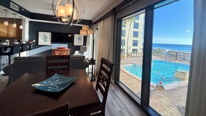 A modern hotel room with a dining area, a kitchen, and a large window overlooking a pool and ocean. - Chic Condo w/ Poolside Balcony & Gulf Views. Free Golf & Daily Activities! (Panama City Beach)