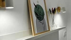 Una obra de arte enmarco con diseño abstracto, un florero con flores secas y dos lámparas fijadas a una repisa blanca.