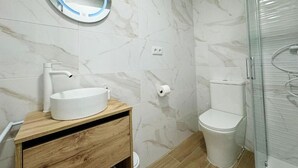 Un baño con pared de mármol, un espejo redondo, un lavabo blanco, un inodoro y una cabina de ducha con mampara de vidrio.