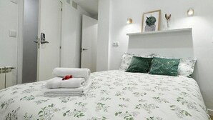 Un dormitorio con cama, paredes blancas, una puerta y un estante con objetos decorativos.