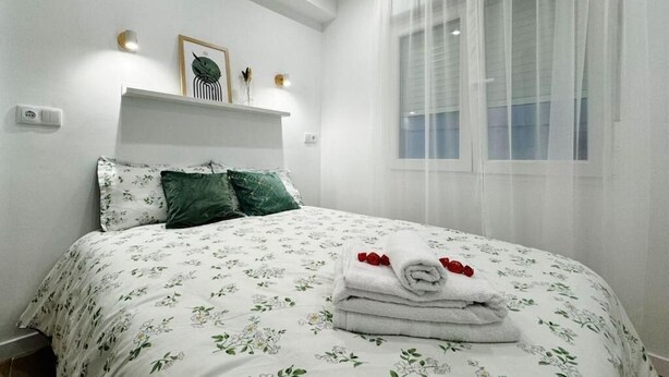 Una cama bien tendida con ropa de cama floral, una mesita de noche con adornos y una ventana con cortinas traslúcidas.