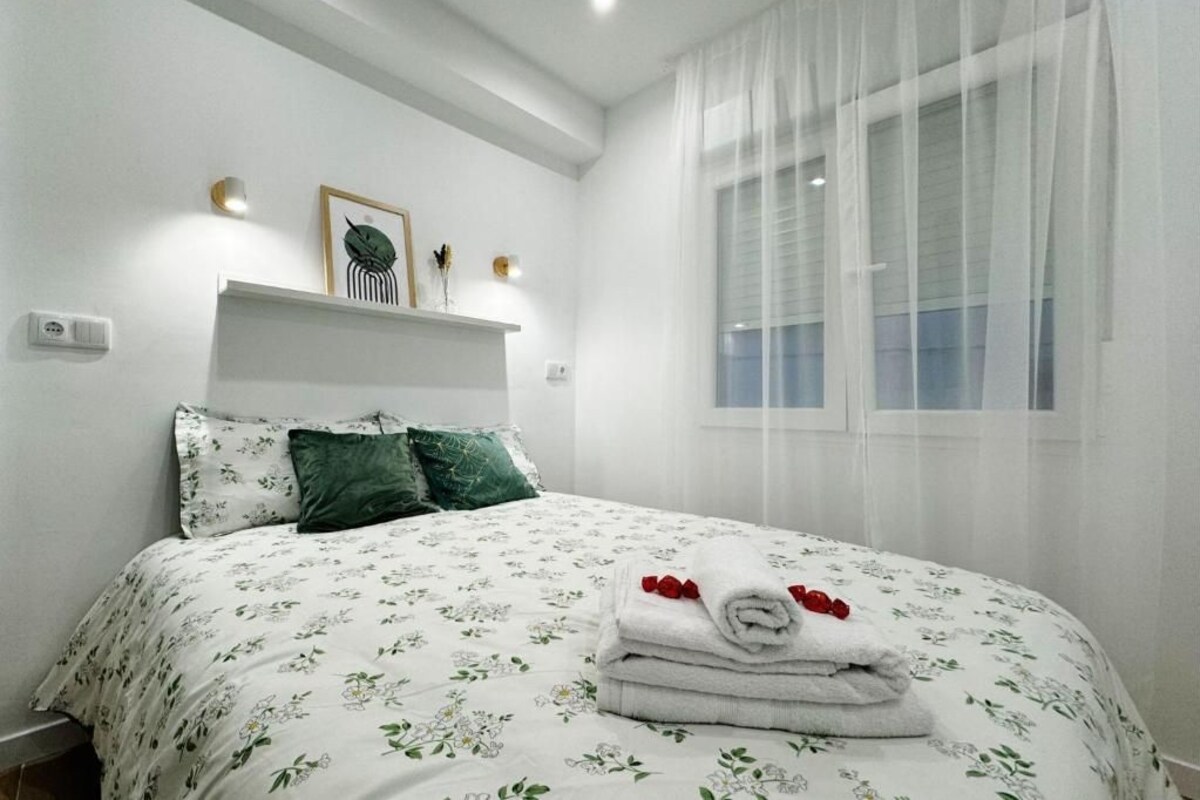 Una cama bien tendida con ropa de cama floral, una mesita de noche con adornos y una ventana con cortinas traslúcidas.