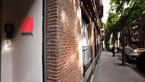 A hostel with a red logo, brick facade, and a narrow sidewalk with parked cars and trees. - Fit Hostel Madrid (Madrid)