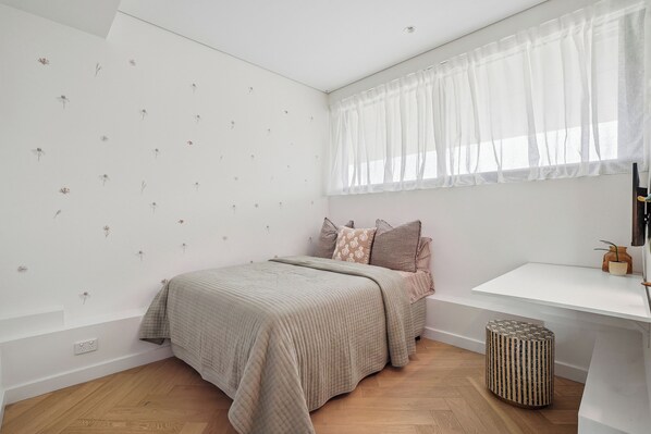 A bedroom with a bed, a bedside table, a window with sheer curtains, and a wall with a floral decal. - Three-Bedroom Ocean-View (Tamarama)