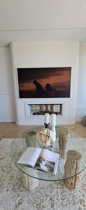 A living room with a fireplace, a large TV mounted above it, a glass coffee table with a book and decorative items, and a patterned rug. - Three-Bedroom Ocean-View (Tamarama)