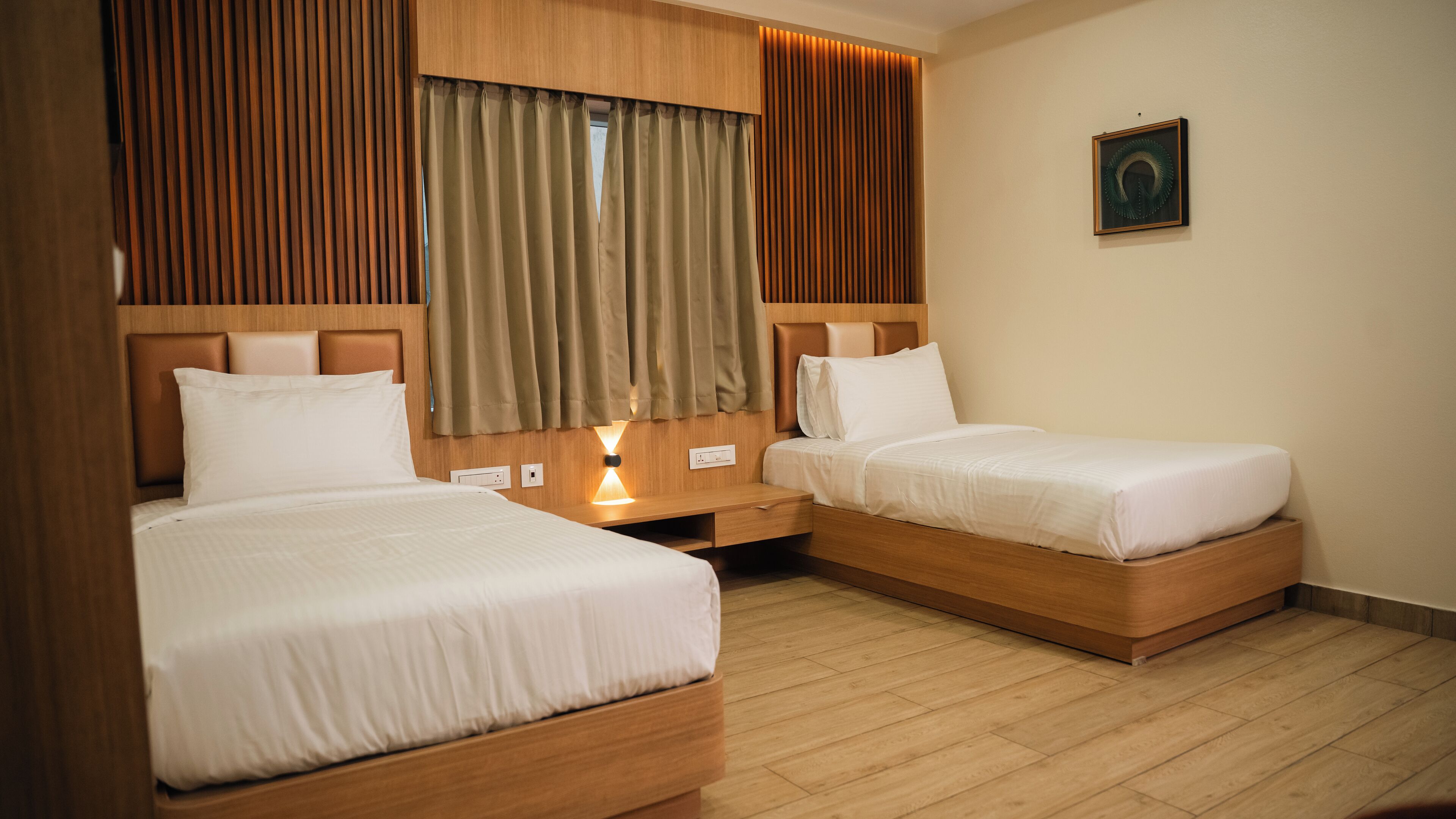 A hotel room with two single beds, wooden headboards, a nightstand with a lamp, and a painting on the wall.