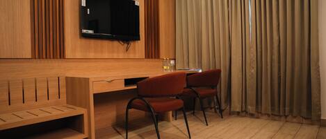 A hotel room with a wooden desk, two red chairs, a television, and a window with curtains.