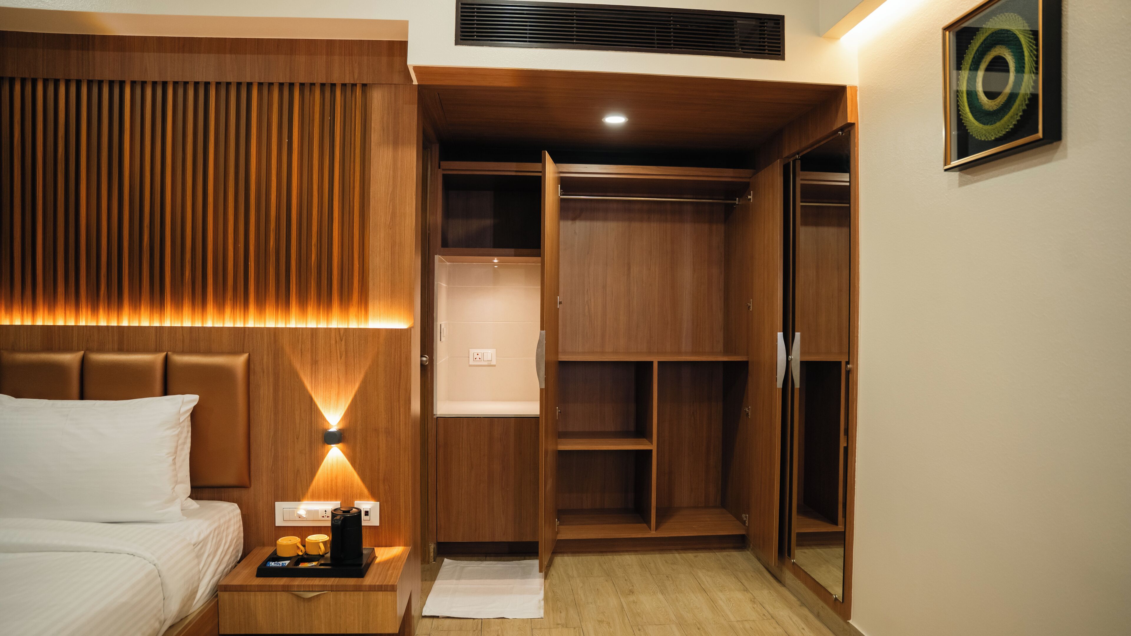 A hotel room with a bed, bedside table with a coffee maker, and a wooden wardrobe with a mirror.