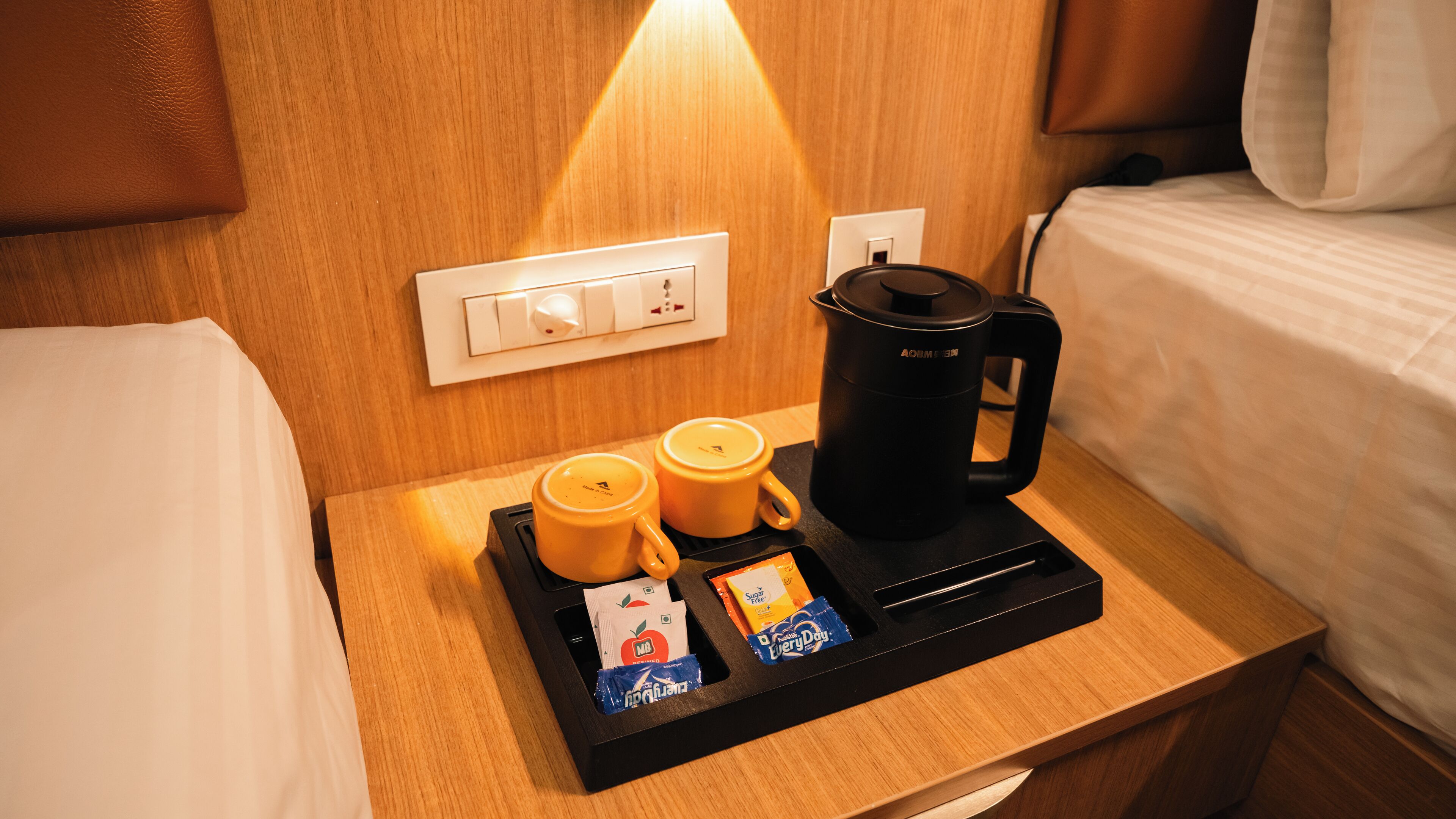 A hotel room with a wooden nightstand, a black coffee maker, two yellow mugs, and a tray with sugar packets and a tea bag.