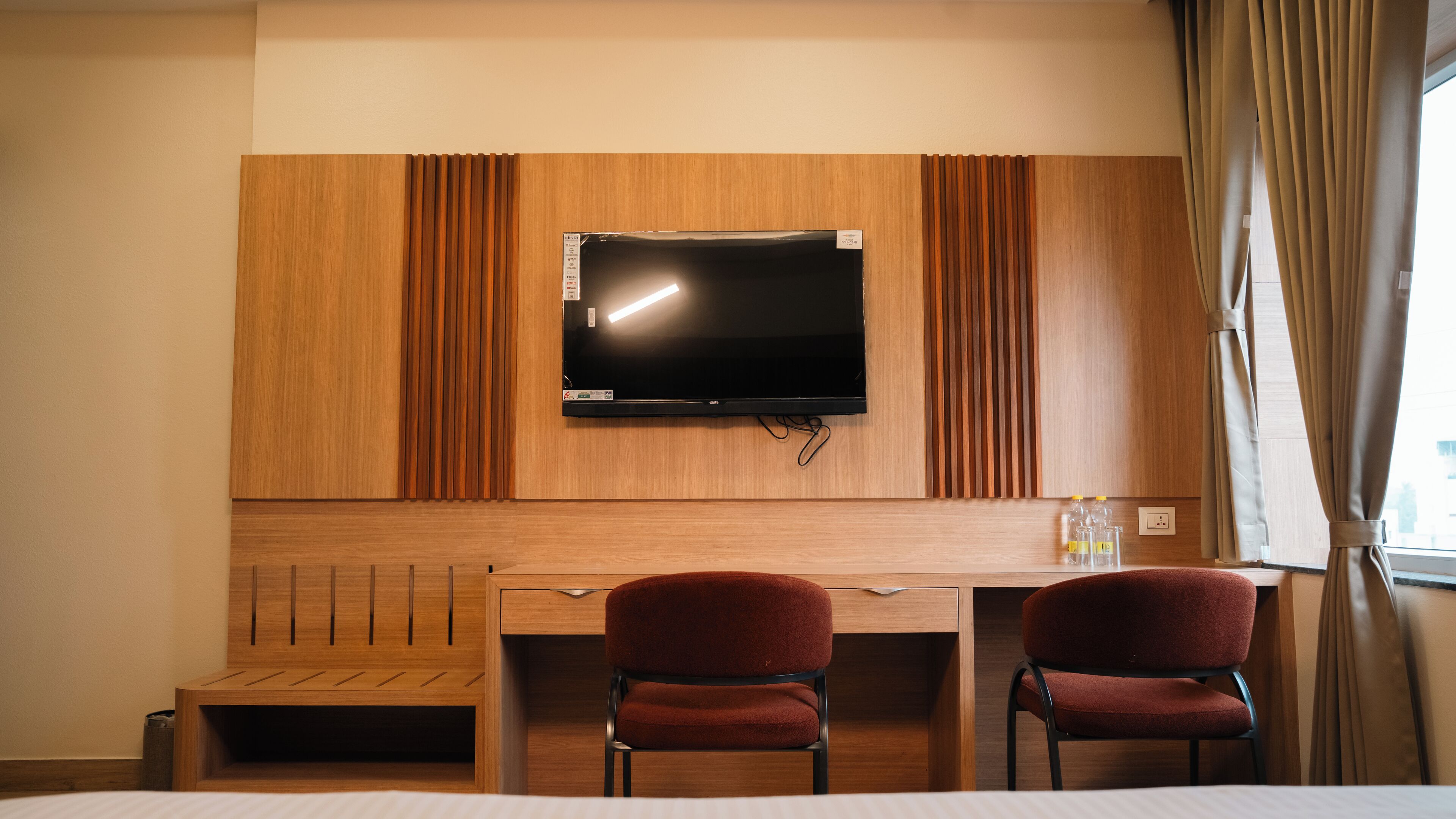 A hotel room with a wooden desk, two red chairs, a flat-screen TV, and a window with curtains.