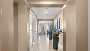 A hallway with beige walls, white trim, and a tiled floor. There are two large potted plants with blue and green foliage. - Penthouse Premium 3BR Condo - 1900+ Sq Ft - Beach Service Included (Panama City Beach)
