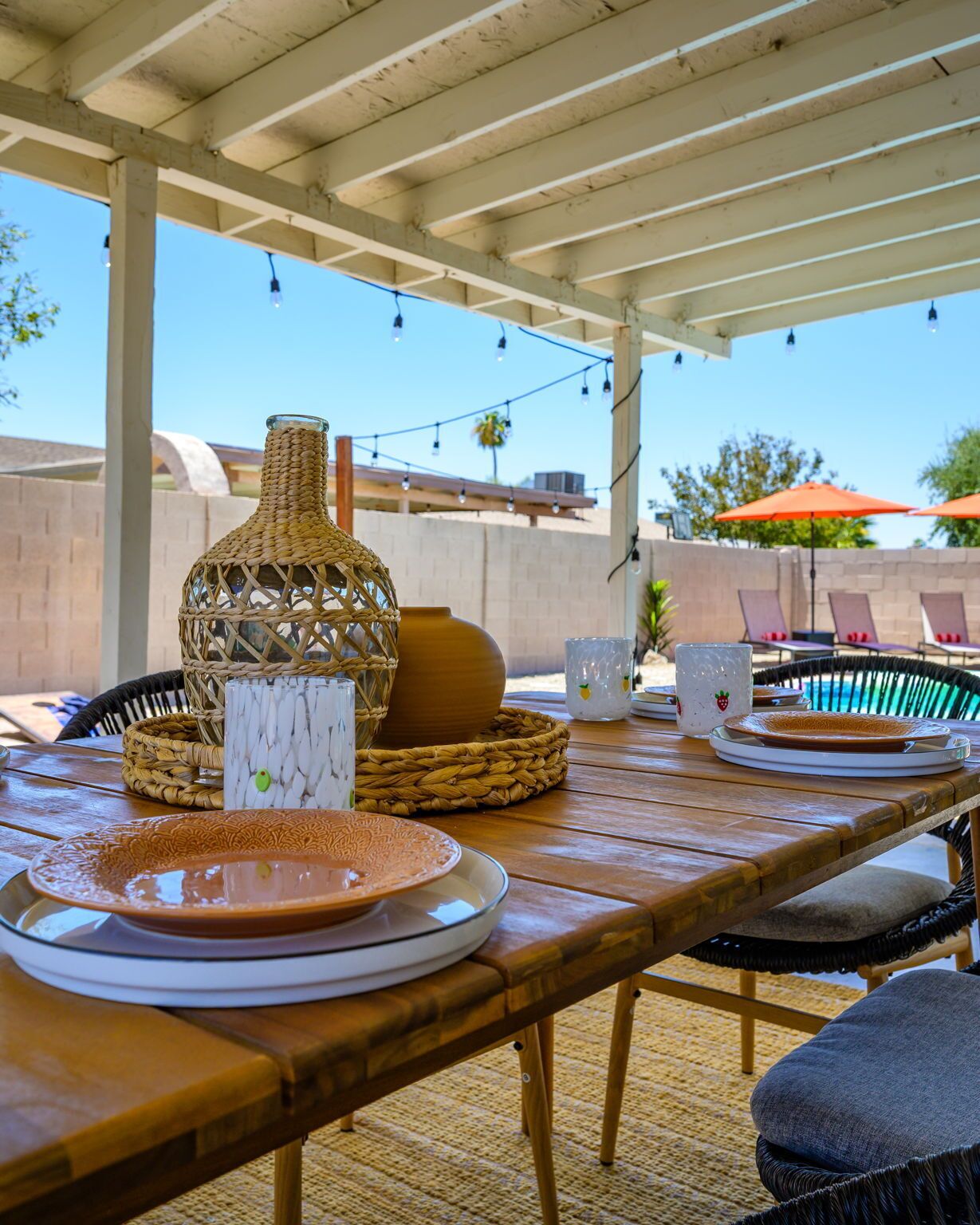 An outdoor patio with a wooden table set for dining, featuring woven baskets, plates, and a decorative vase.