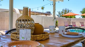An outdoor patio with a wooden table set for dining, featuring woven baskets, plates, and a decorative vase.
