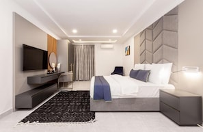 A modern bedroom with a large bed, a TV, a bedside table, and a built-in headboard. - STP Apartments (Abuja)